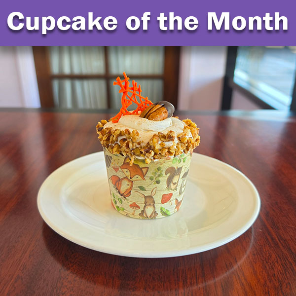 A decorated cupcake displayed on a white plate atop a wooden table. The cupcake features a critter-themed wrapper with autumn colors, frosted with cream-colored frosting topped with crushed nuts or cookie crumble, and garnished with a bright orange carrot decoration