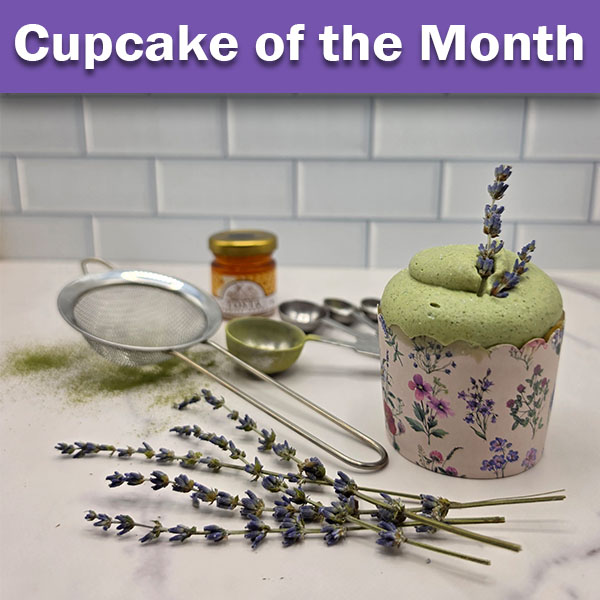 A single green matcha cupcake in a purple floral wrapper, topped with lavender sprigs. The scene is set with a background of a small honey jar, measuring spoons, a fine mesh sieve, scattered green powder, and more fresh lavender stalks on a white surface.