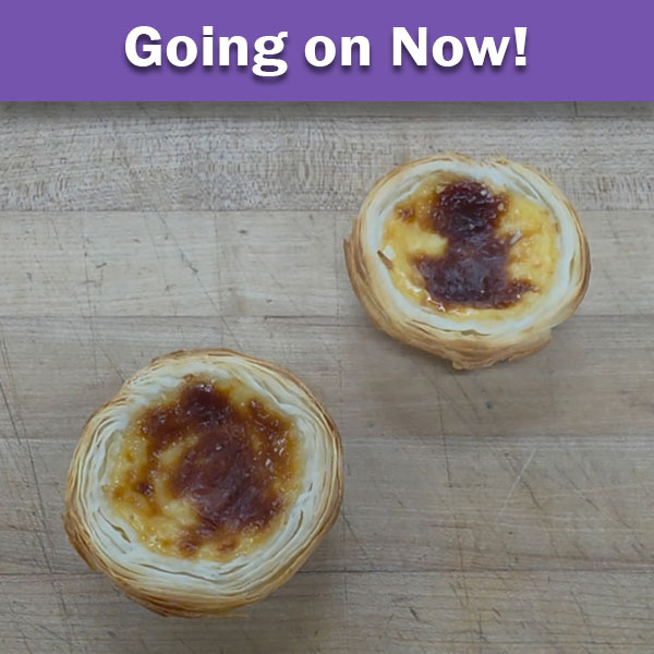A top-down view of two Portuguese custard tarts Pastel de Natas (Pasteis de Nata) with flaky, golden-brown crusts and caramelized tops, resting on a worn wooden butcher block surface.